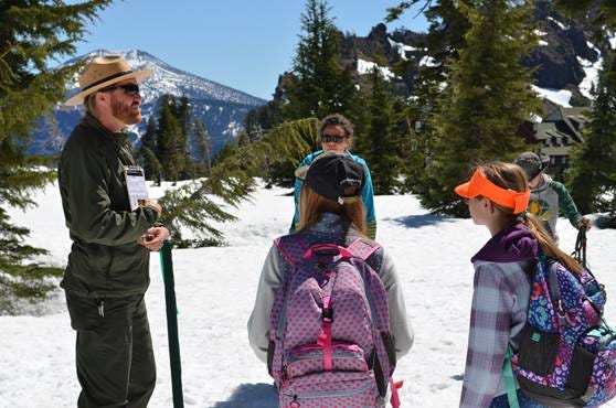A man wearing a park ranger's hat stands on a snow-topped hill talking to a group of four children